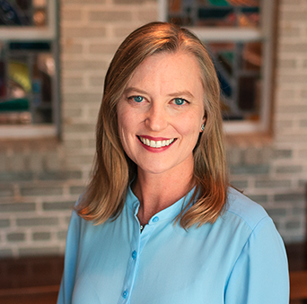 A woman in a blue shirt is smiling in front of a brick wall.