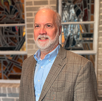 A man in a suit and blue shirt is standing in front of a stained glass window.