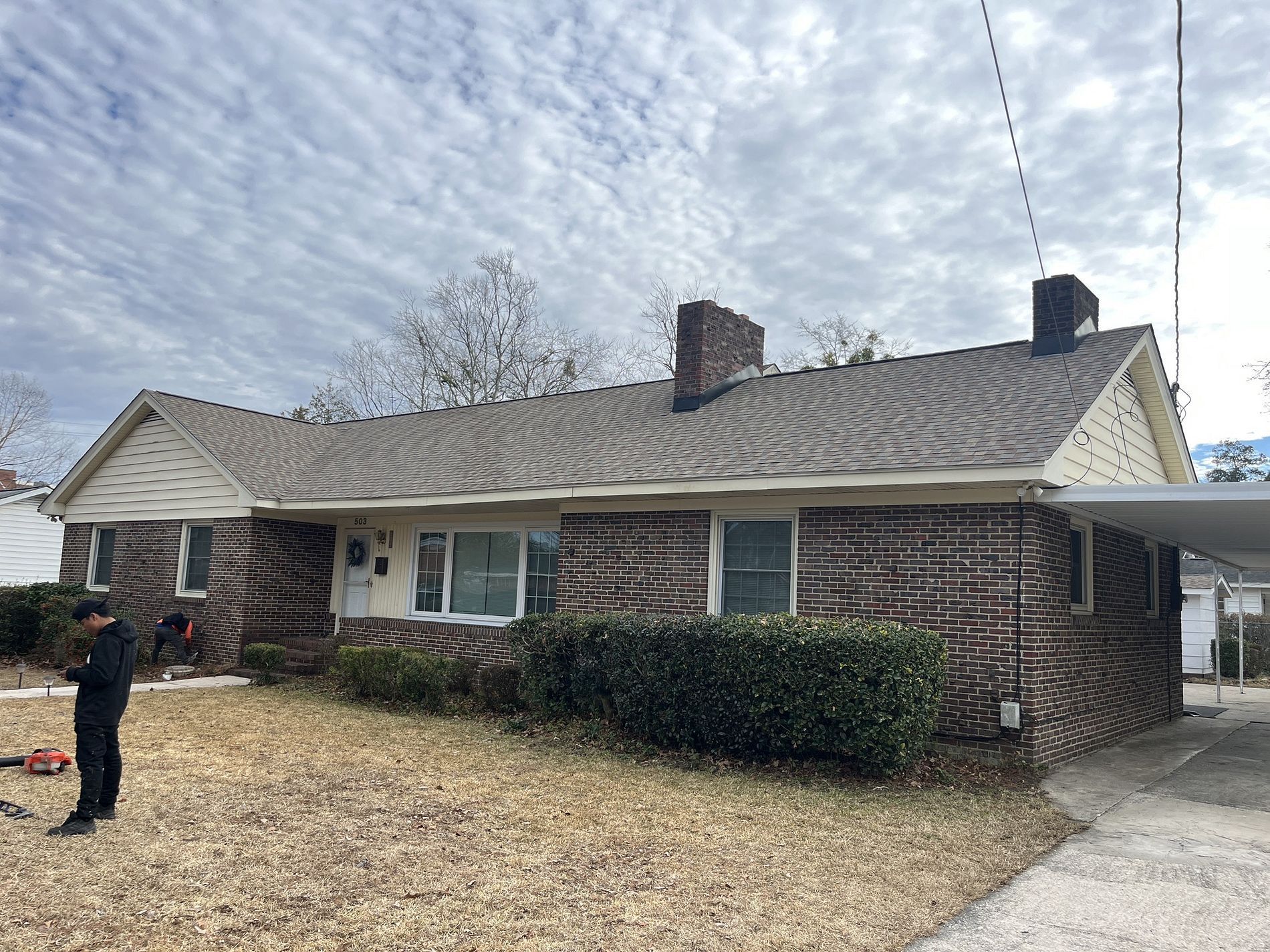 A single-story brick house with a new grey shingled roof, two chimneys, and a covered carport, under a cloudy sky.