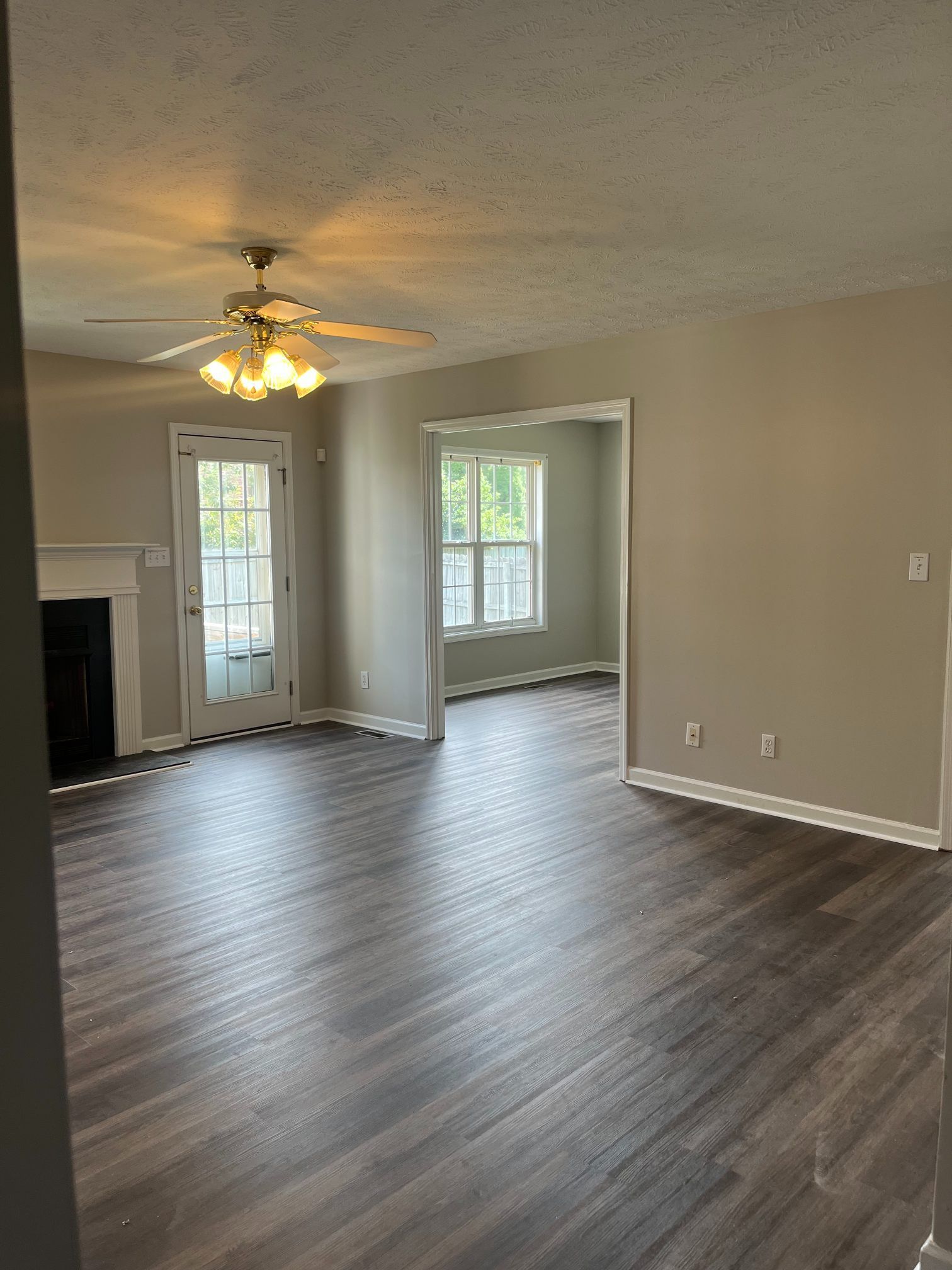 Interior of a living room with dark wood-look flooring and neutral walls. A doorway, windows, and fireplace are visible.