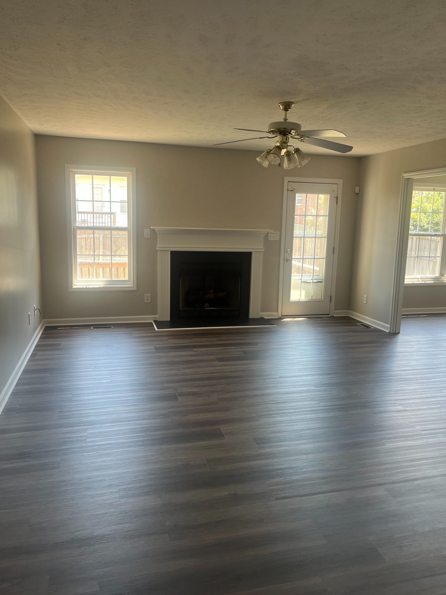 Empty living room with dark wood-look floor, fireplace, windows, and a ceiling fan.