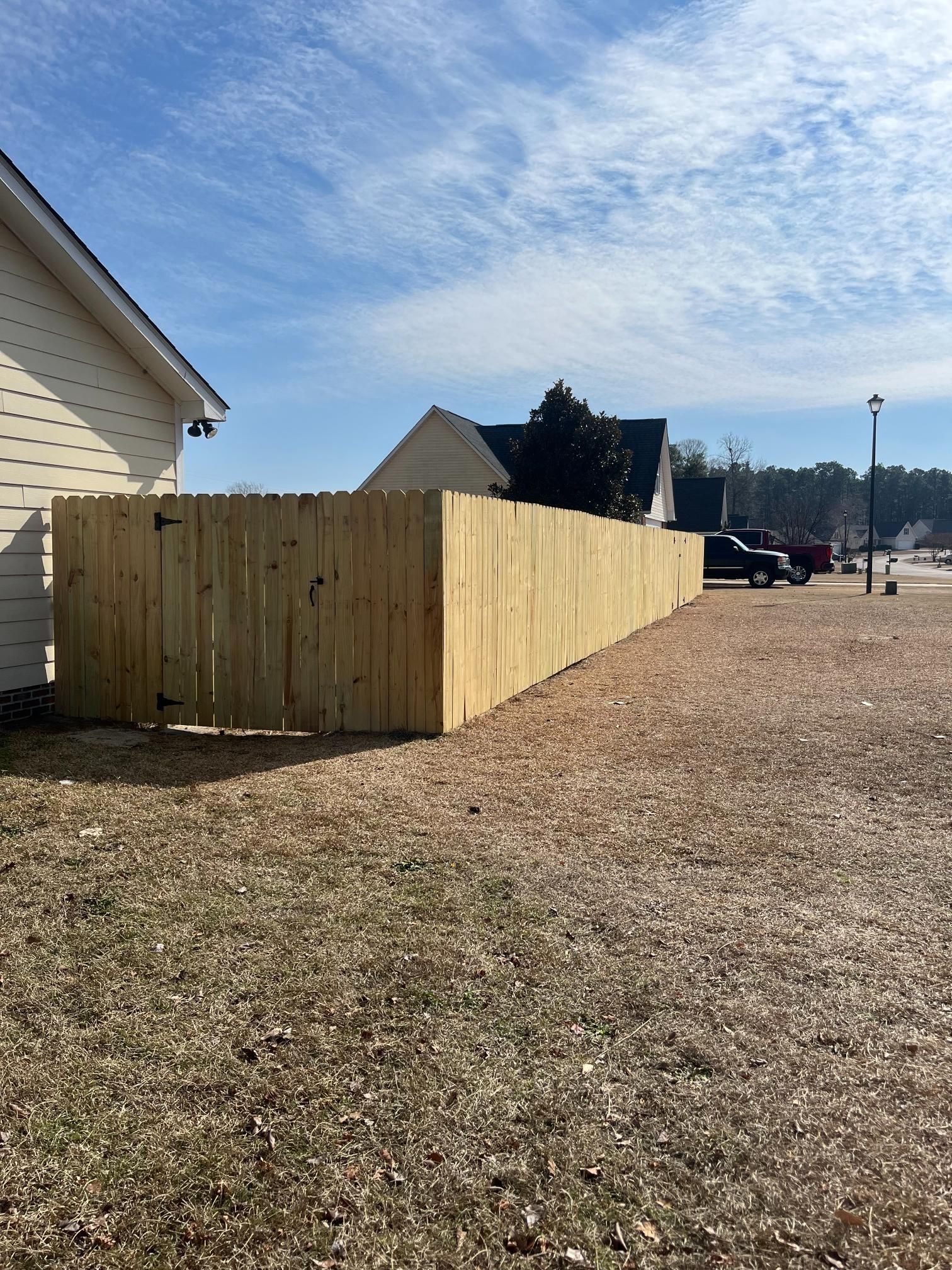 Wooden fence along a gravel area in front of houses under a partly cloudy blue sky.