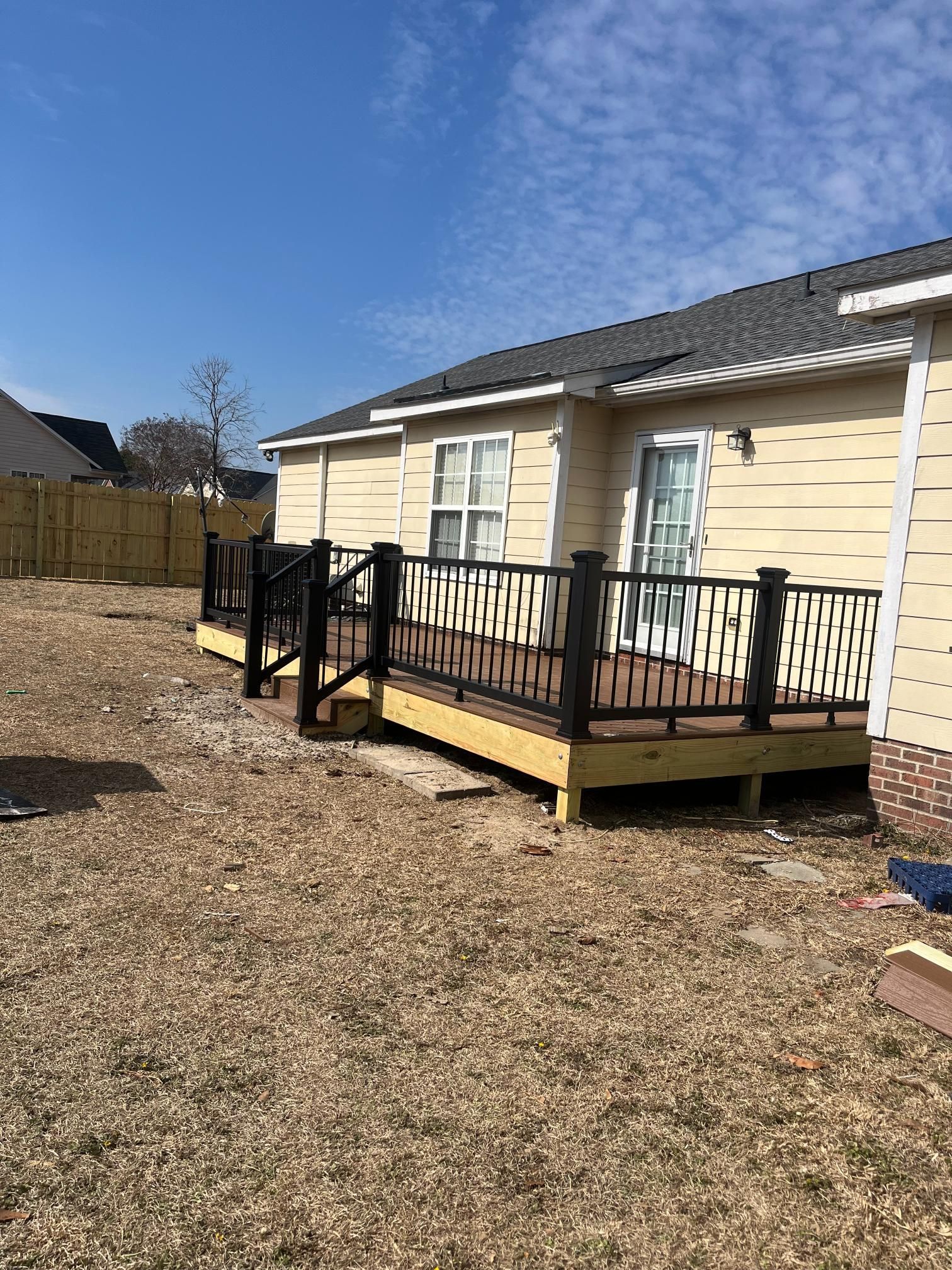 Newly constructed wooden deck with black railings next to a light yellow house, with a fence in the background.