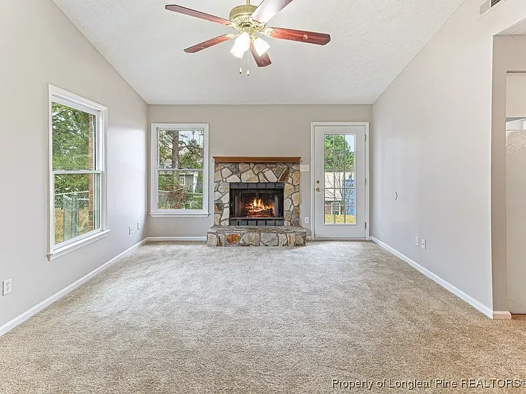 Empty living room with fireplace, windows, and door; light carpet and walls.