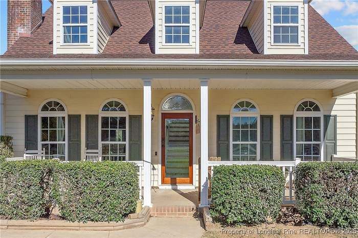 A light yellow one-story house with a front porch, three roof dormers, green shutters, and trimmed hedges in front.
