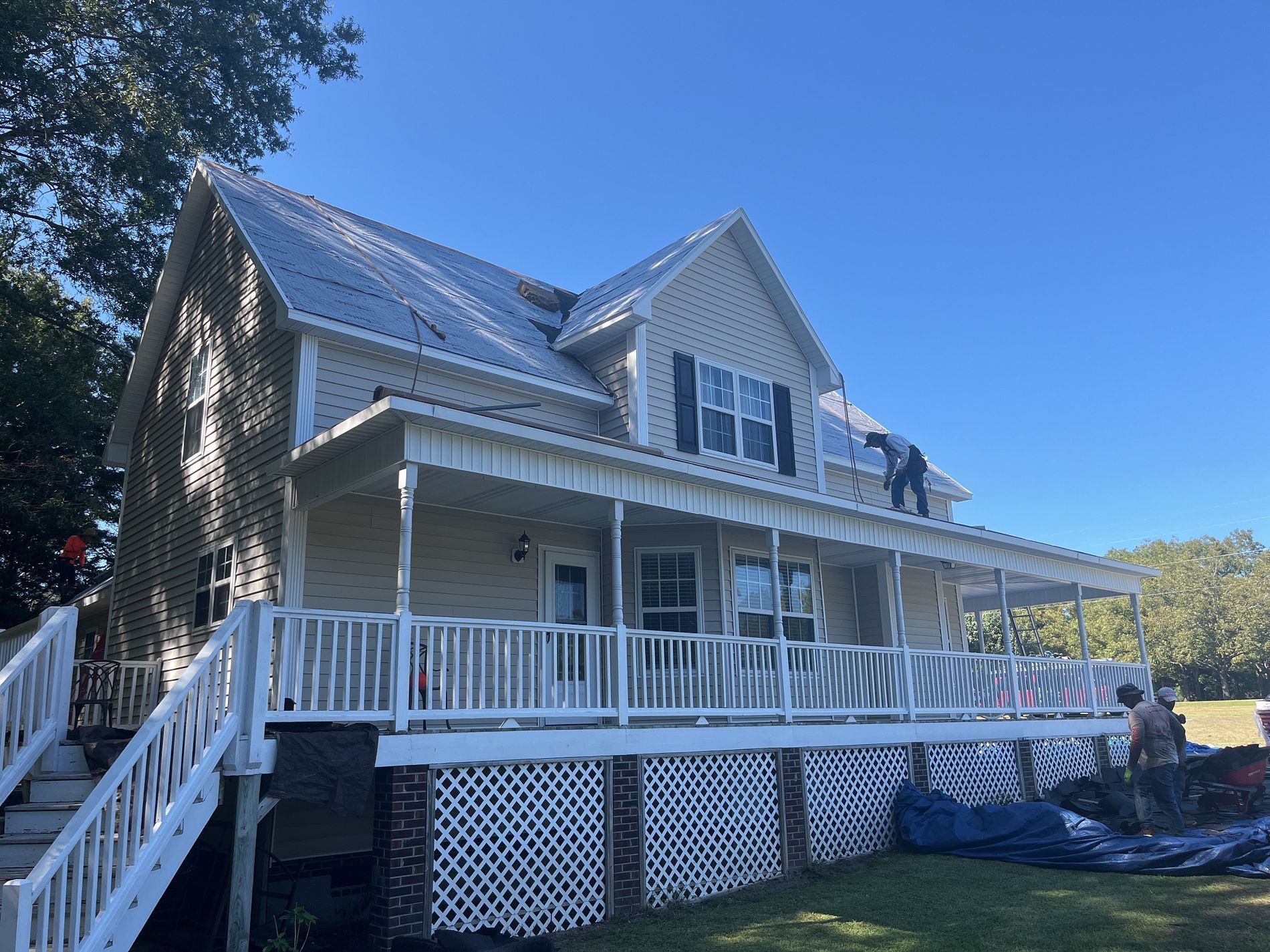 Two-story beige house with white porch and metal roof under a blue sky, workers on roof.