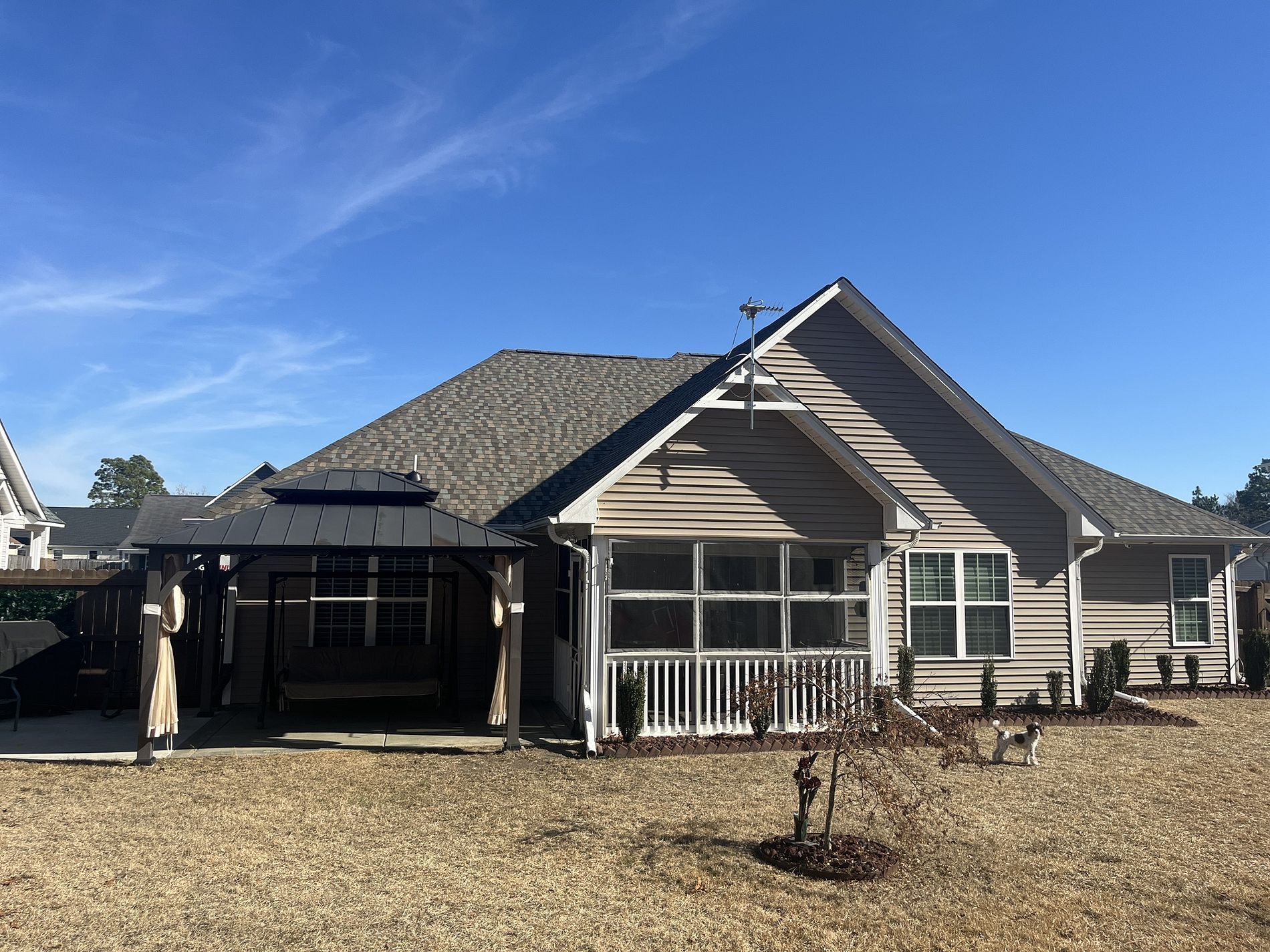 The rear of a single-story beige house with a screened porch, an attached metal gazebo, and a gravel yard under a blue sky.