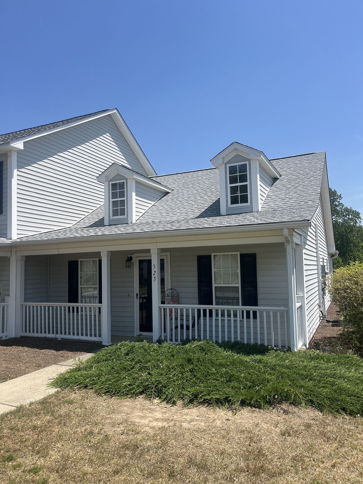 Light gray house exterior with a covered porch, white railings, black shutters, and two dormer windows under a blue sky.