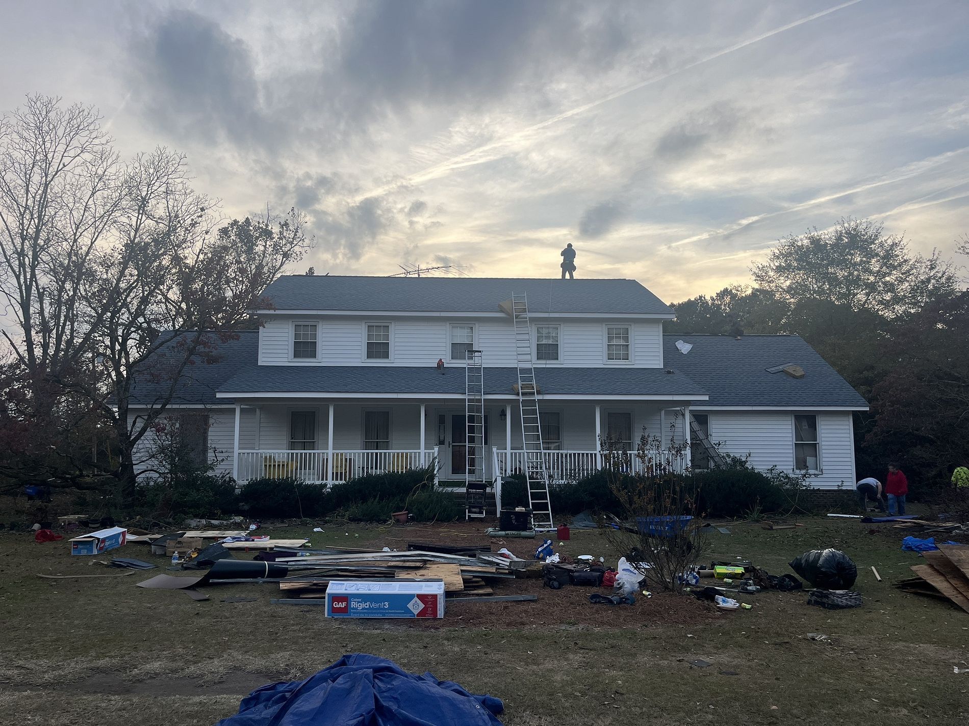 A white house with a partially replaced gray roof, a ladder against the porch, and debris scattered across the front yard.