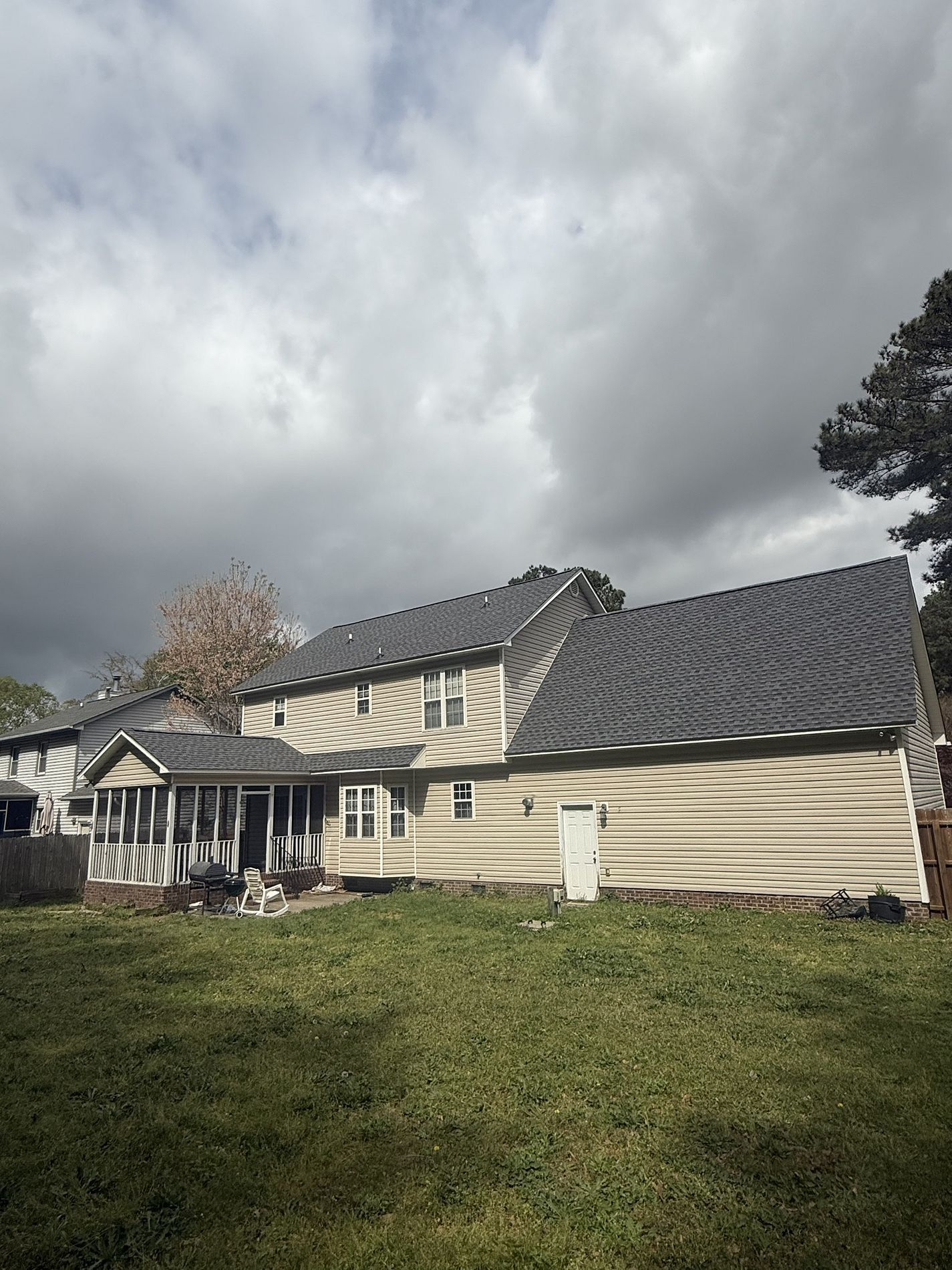Backyard view of a two-story beige house with a dark gray shingled roof and a screened-in porch under a cloudy sky.