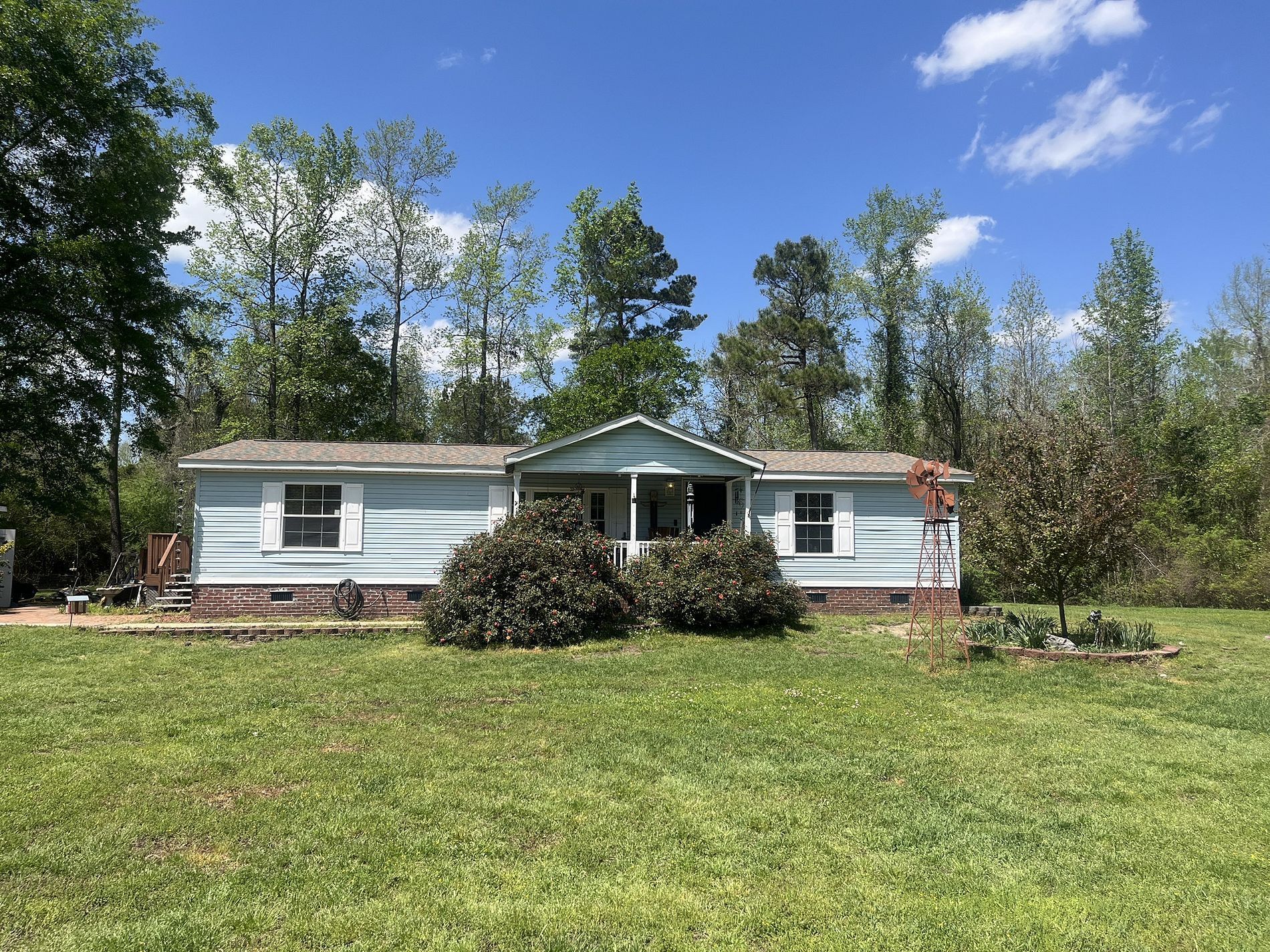A light blue single-wide mobile home with a small front porch, situated on a grassy lot surrounded by trees.