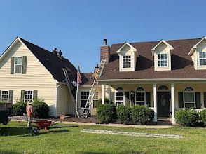 Workers on a roof, ladder leaning against the house. Sunny day with green grass and beige house.