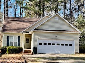 Tan house with a brown roof and a two-car garage. A front door and shrubs are visible.