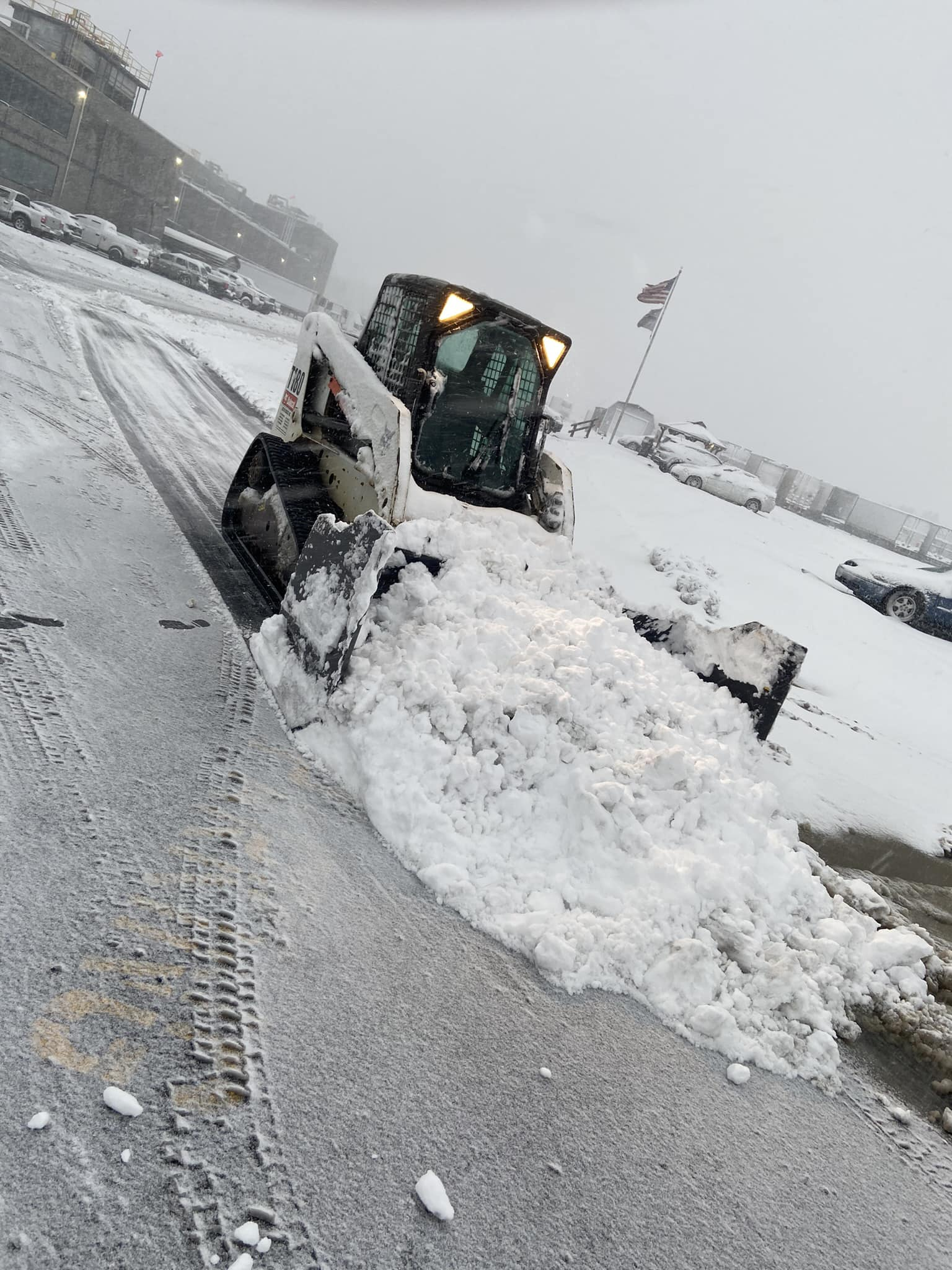 Bobcat clearing snow from a parking lot on a snowy day; buildings visible in the background.