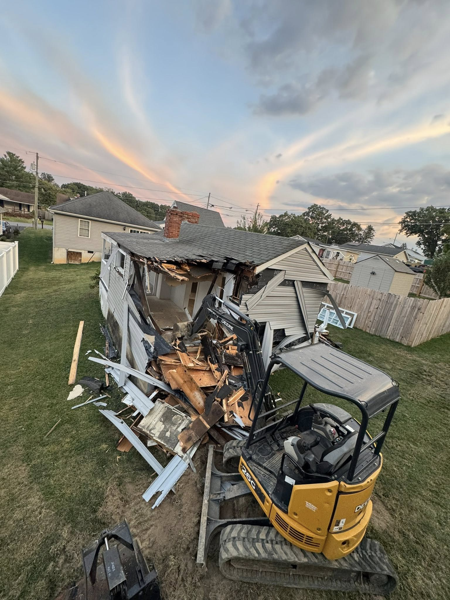 An excavator demolishes a house; wood and debris spread across the grass yard.