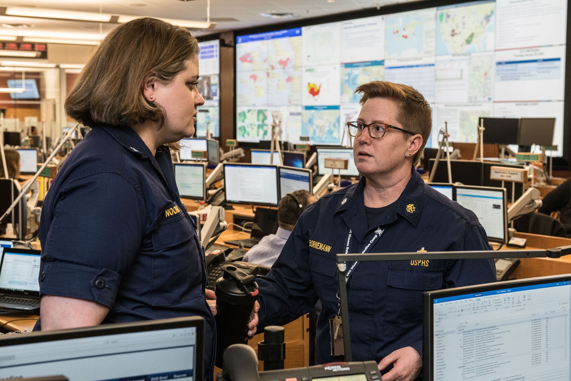 Two people in navy uniforms talking in a busy communications center with monitors and wall maps.