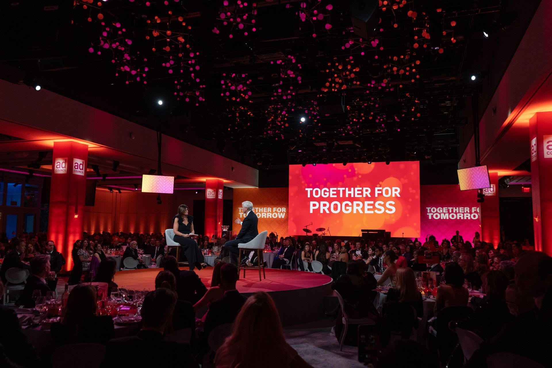 A woman is sitting on a stage in front of a large screen that says together for progress.
