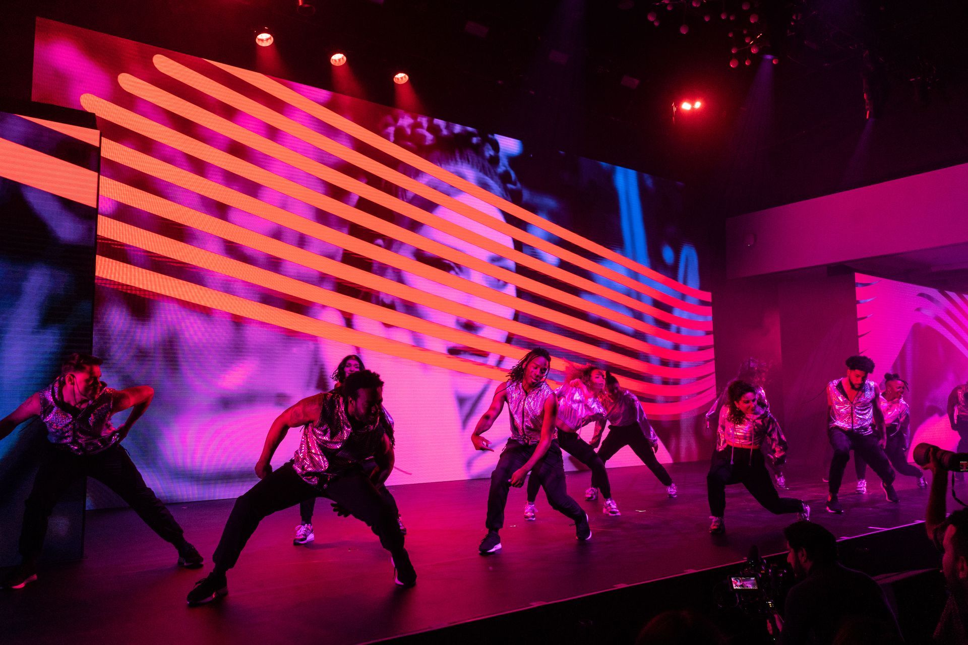 A group of people are dancing on a stage in front of a large screen.