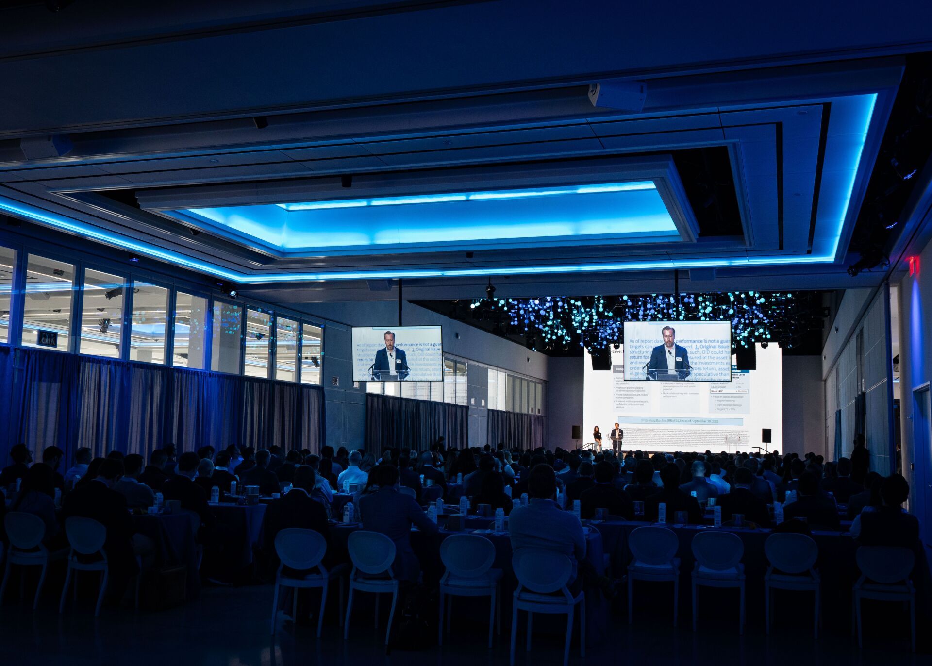 A large room with a lot of people sitting at tables in front of a large screen.
