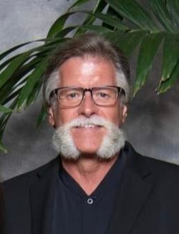 A man with glasses and a beard is standing in front of a palm tree.