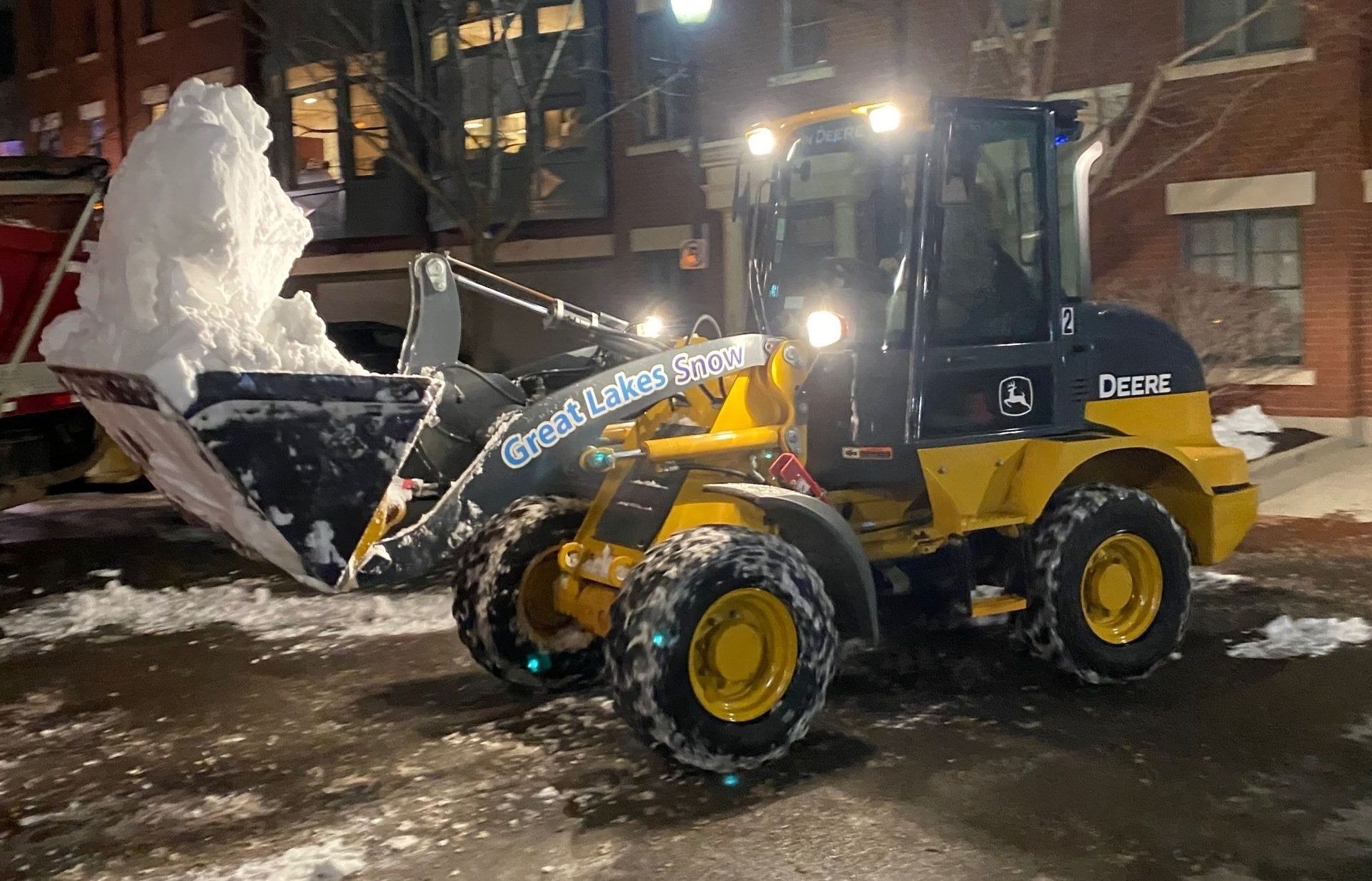 A yellow and black tractor from Great Lakes Snow is clearing snow from a parking lot in Woodstock, IL