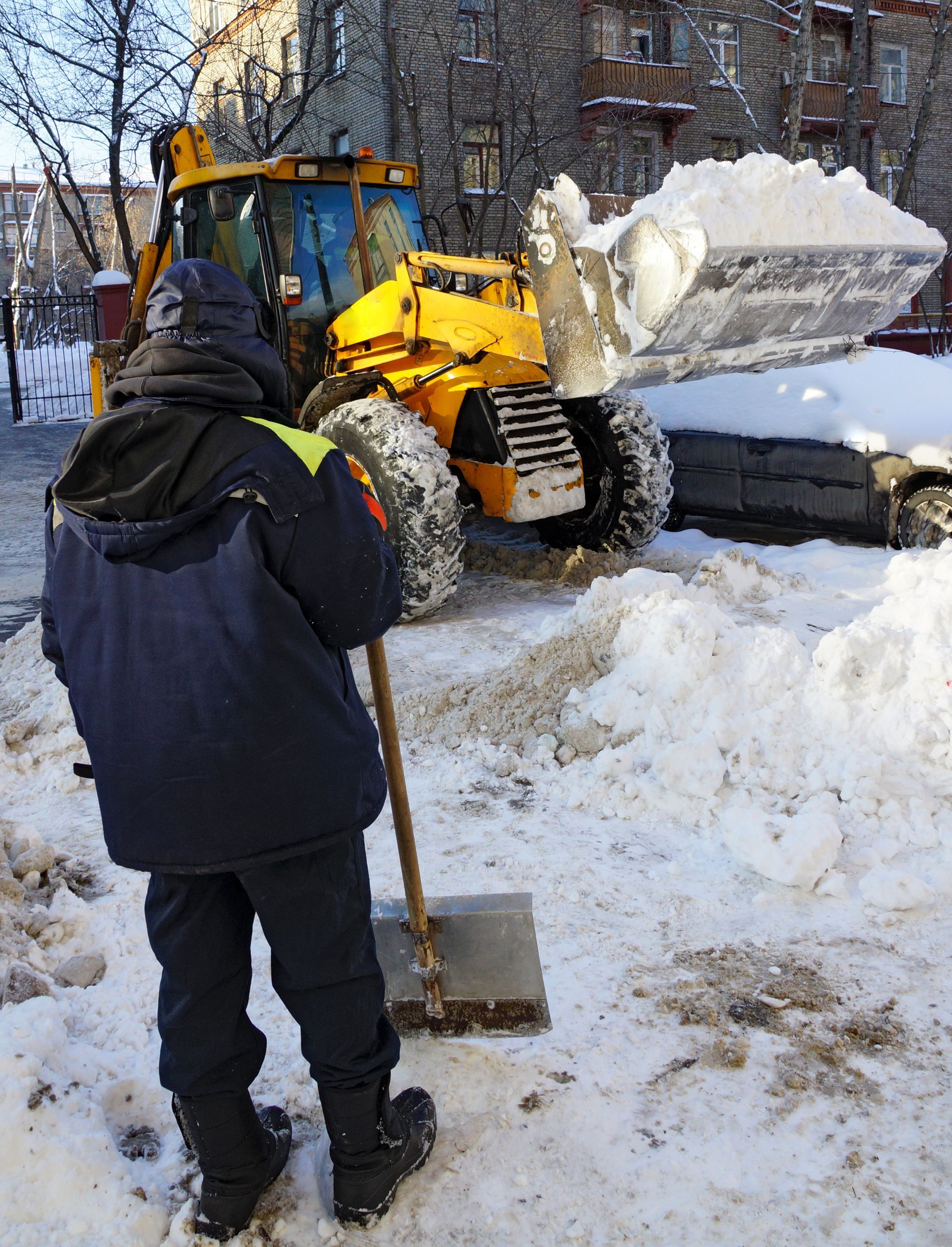 a man is shoveling snow in front of a yellow tractor in front of an apartment building for professional commercial snow and ice removal