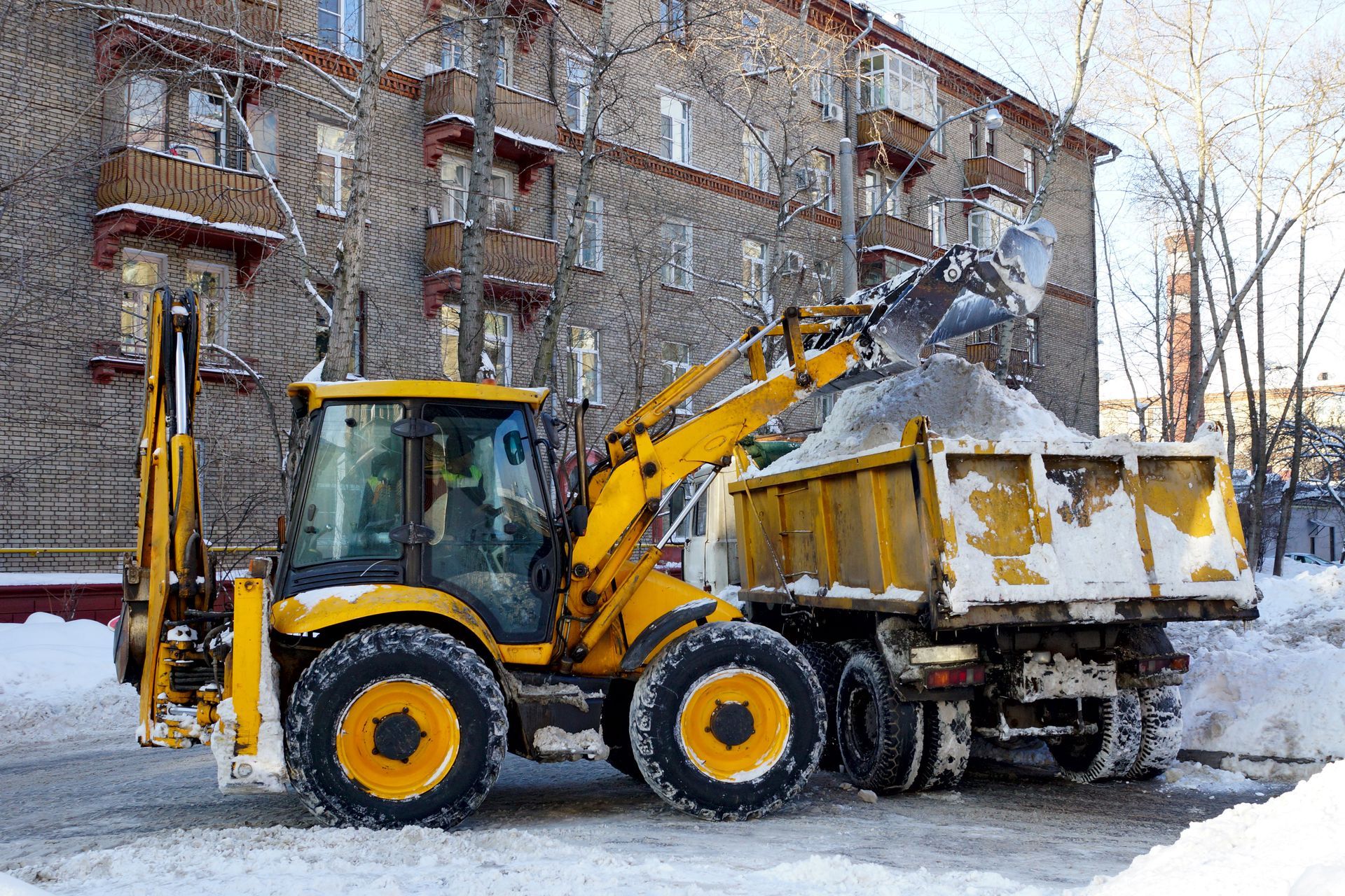 a yellow tractor is loading snow into a dump truck in front of an apartment building -- commercial snow removal