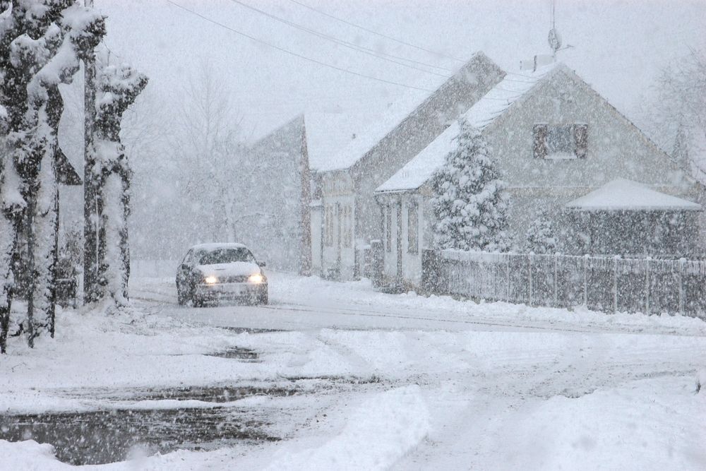 Snowy street scene with a car driving past houses. Heavy snowfall obscures visibility.