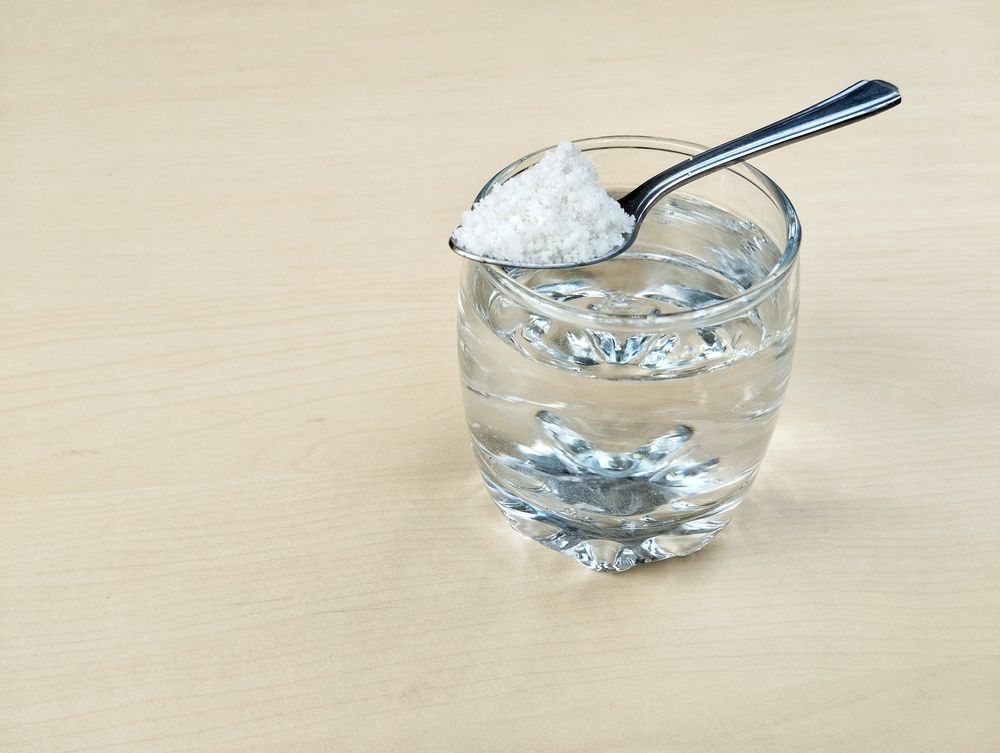 Spoonful of salt resting on the rim of a water-filled glass on a light wood surface.