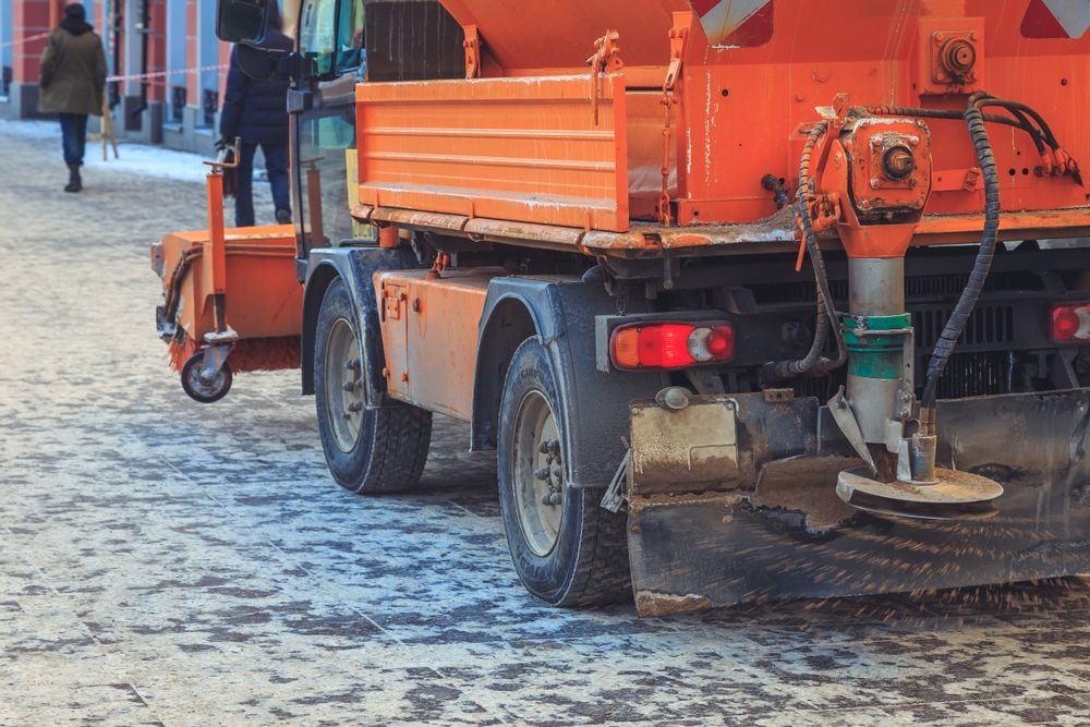 Orange road-sweeping truck cleaning a snowy street with a worker nearby