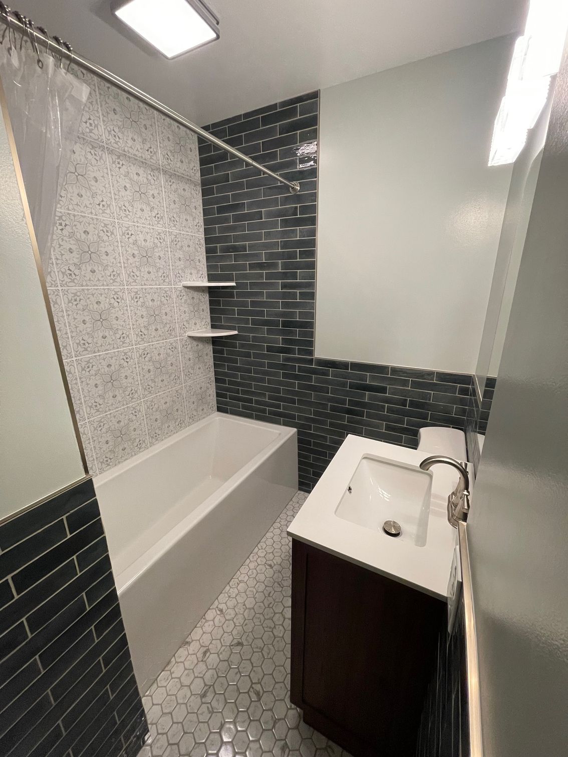 Bathroom with white tub, dark blue-tiled wall, white sink on a brown cabinet, and a gray-tiled floor.