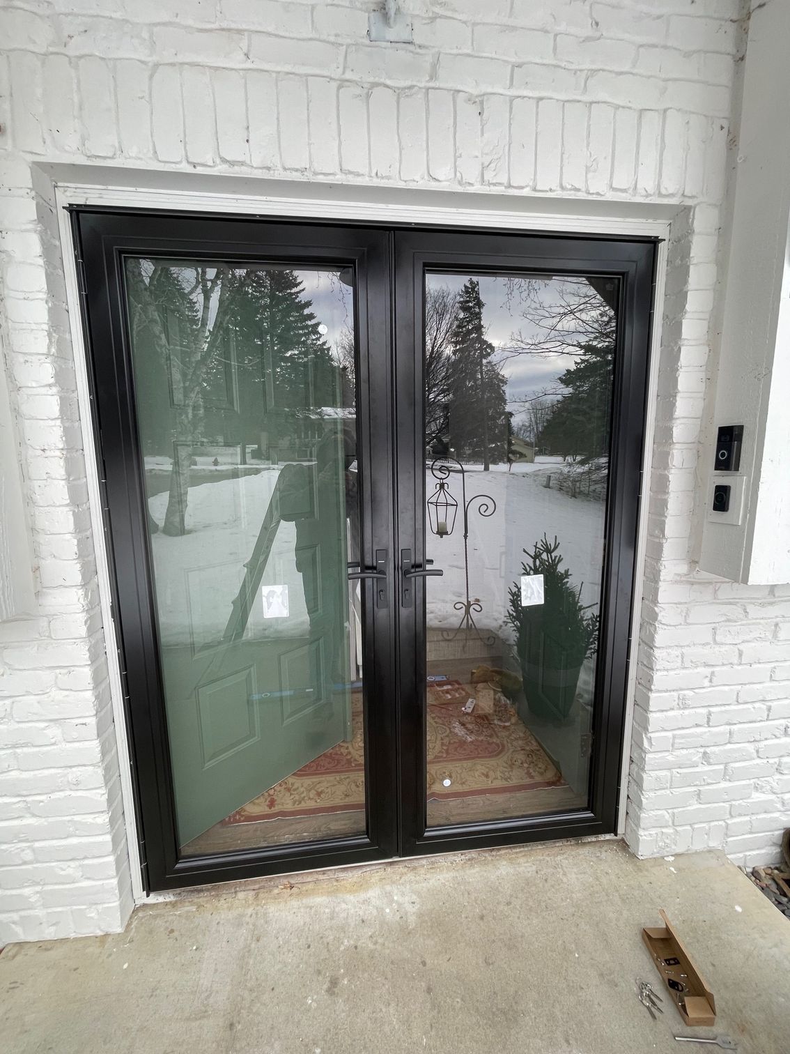 Black-framed glass double doors in a white brick entranceway; one door ajar revealing an interior with a green door and rug.