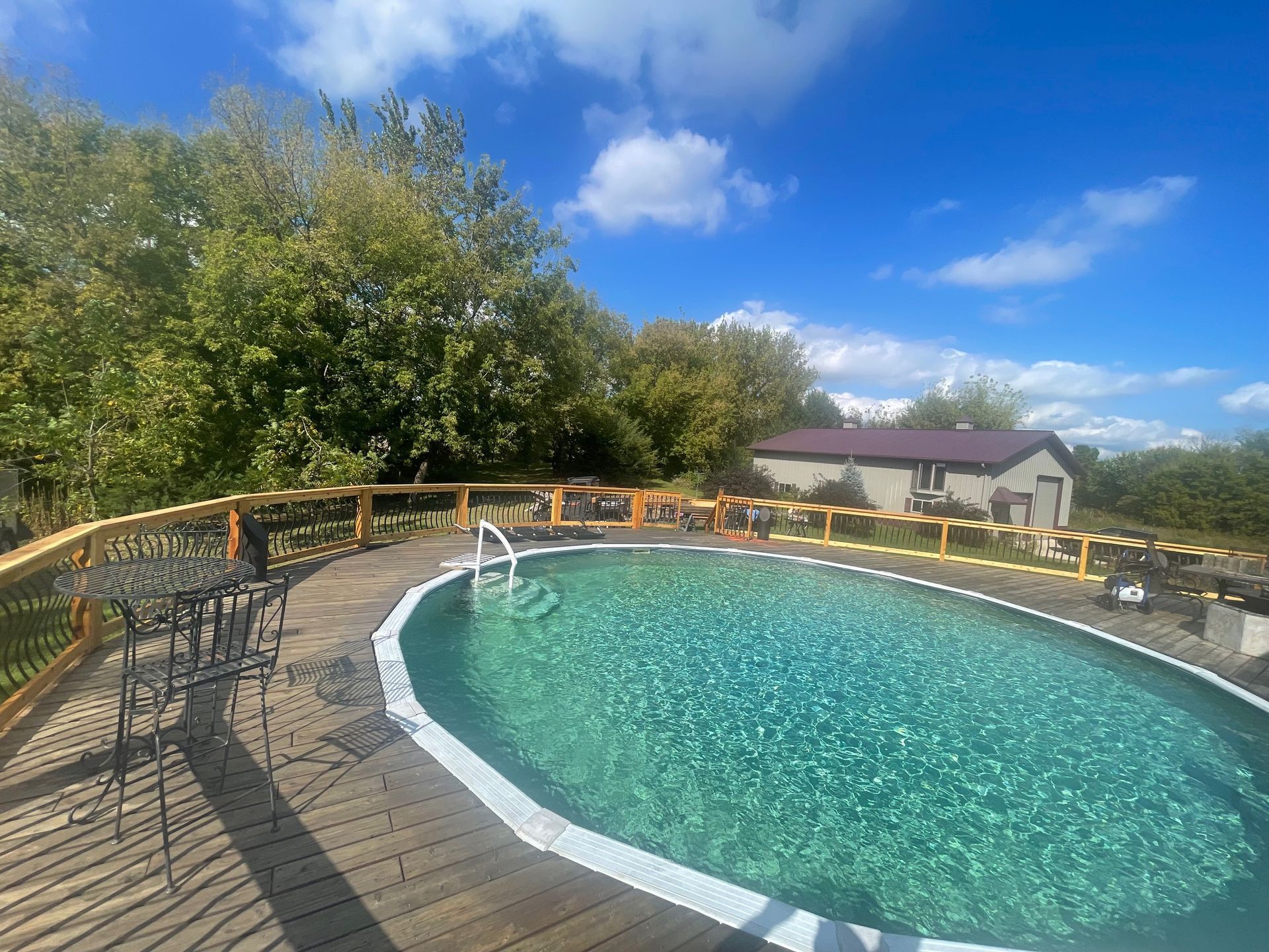 Pool with wooden deck, small building, and trees under a blue sky.