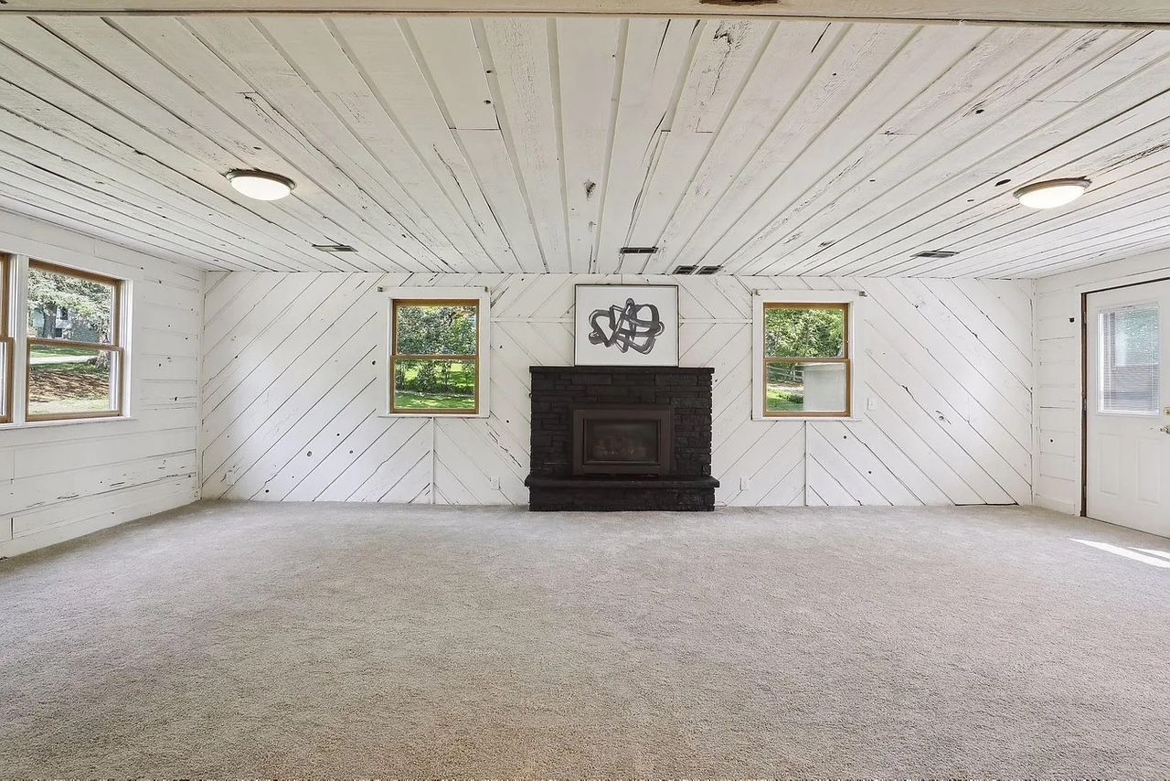 Empty room with white wood walls and ceiling, gray carpet, black fireplace, and two windows.