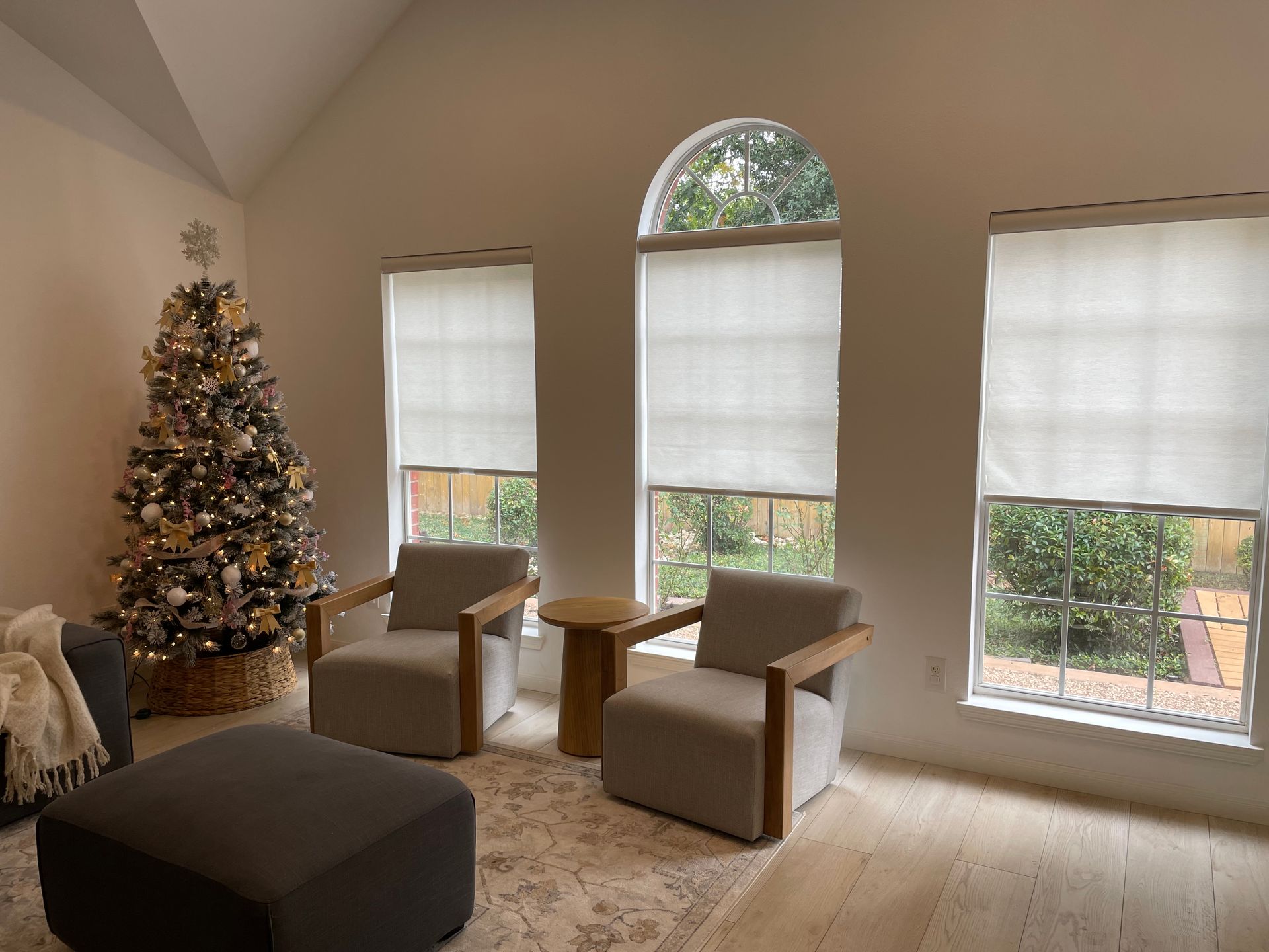 A living room with a christmas tree , chairs , ottoman and a table.