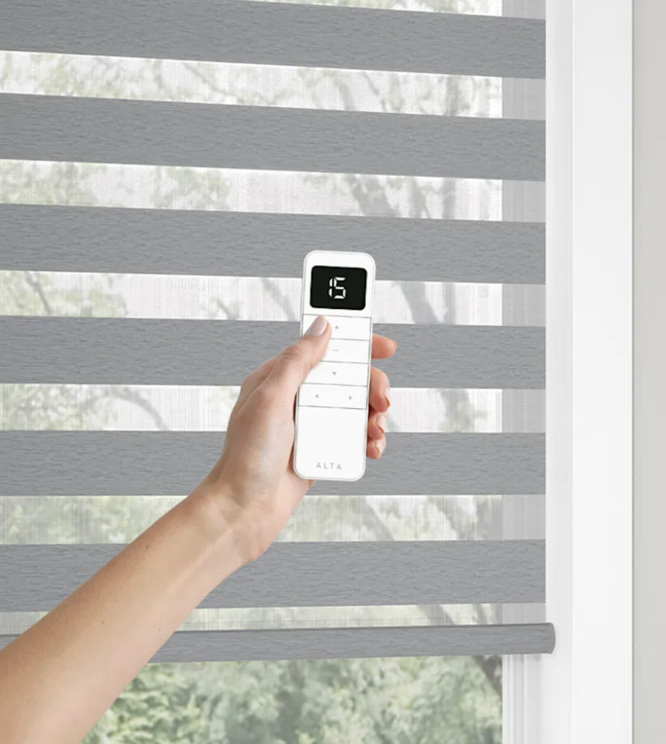 A person is holding a remote control over a window blind.