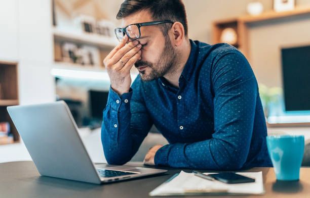 Man with glasses holding his hand to his forehead, looking sleepy while sitting in front of a laptop.