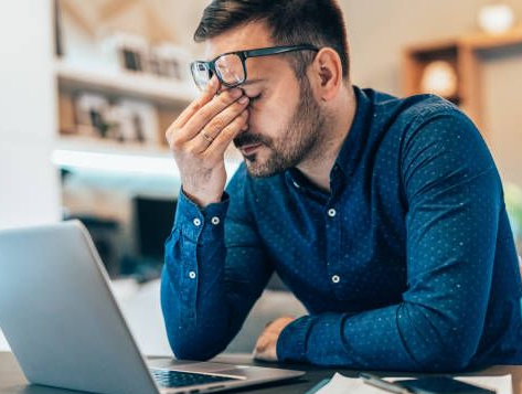 Man with glasses, hand on face, appears stressed while working on laptop.