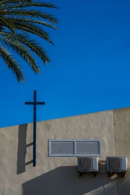 Blue cross on textured wall, palm frond, clear blue sky.