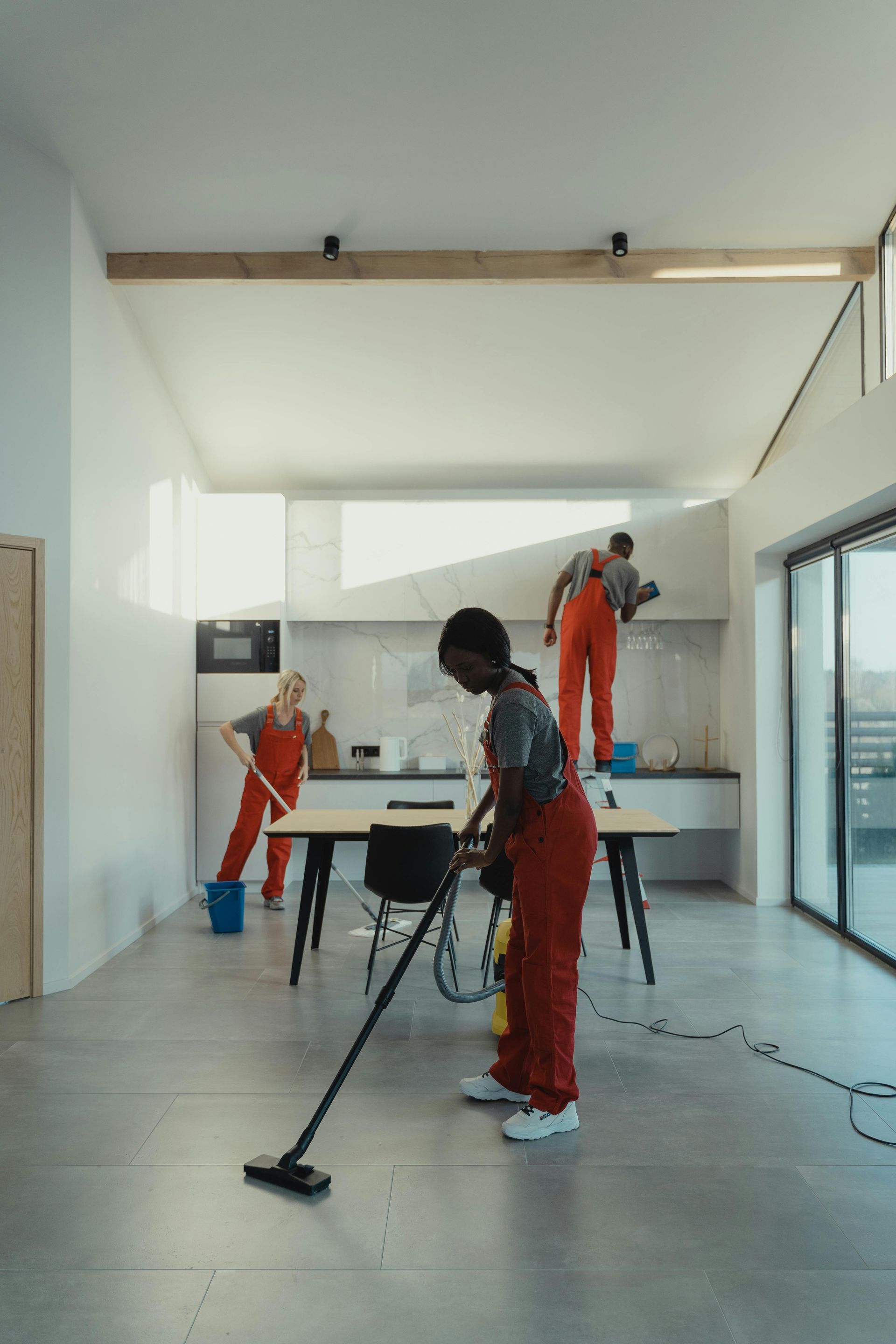 Four cleaning crew members in red jumpsuits cleaning a bright, modern kitchen and living space.