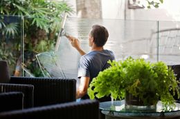 Man cleaning glass with a squeegee; outdoor setting with plants, glass table and black chairs.