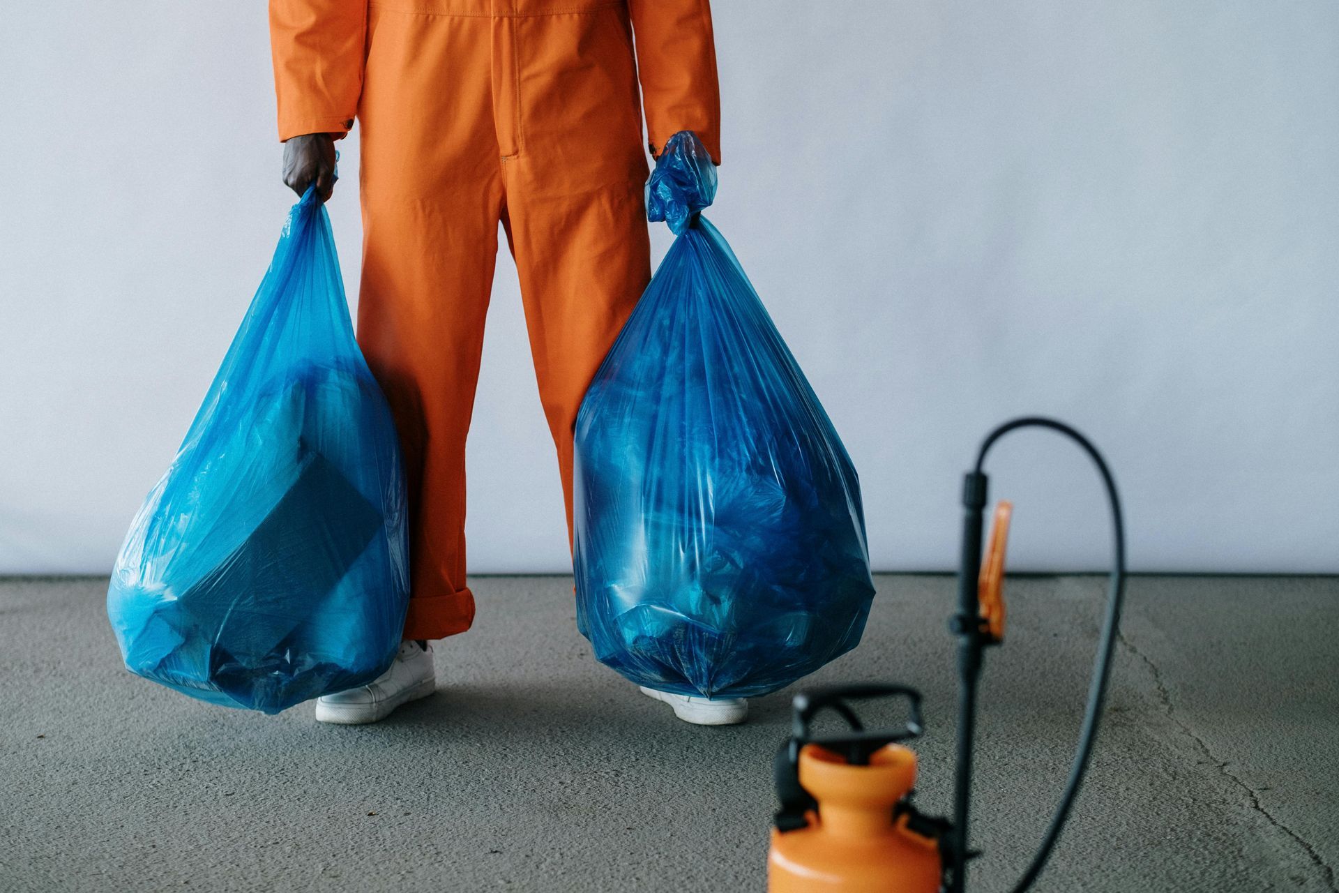 Person in orange jumpsuit holding two blue trash bags, next to a pesticide sprayer.