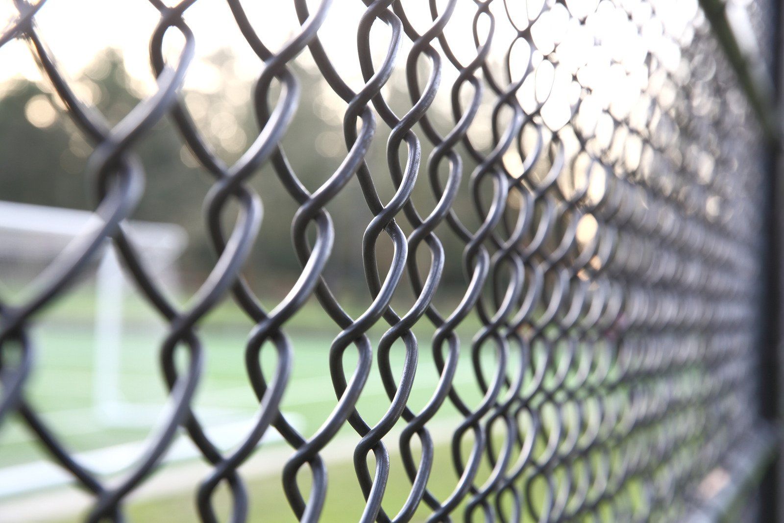 A close up of a chain link fence with a soccer field in the background.