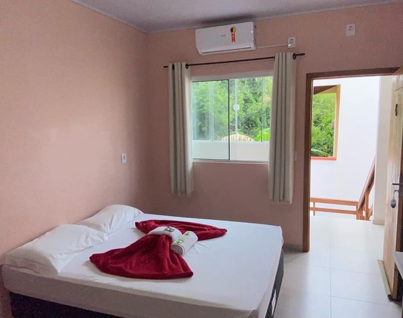 Um quarto arrumado com paredes cor-de-rosa apresenta uma cama com lençóis brancos e uma toalha vermelha dobrada, uma janela e uma porta aberta.