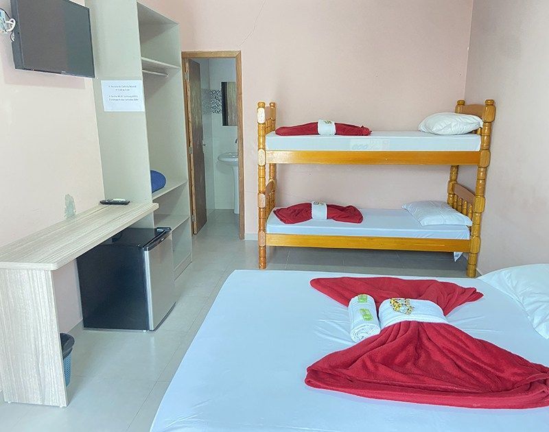 Um quarto de hotel com uma cama grande em primeiro plano, um beliche de madeira ao fundo e uma área de escritório com frigobar.