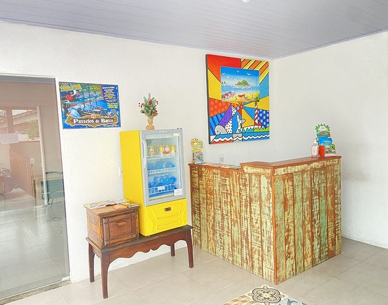 A área de recepção de um hotel apresenta uma escrivaninha rústica de madeira, um frigobar amarelo sobre um suporte de madeira e uma vibrante pintura abstrata.