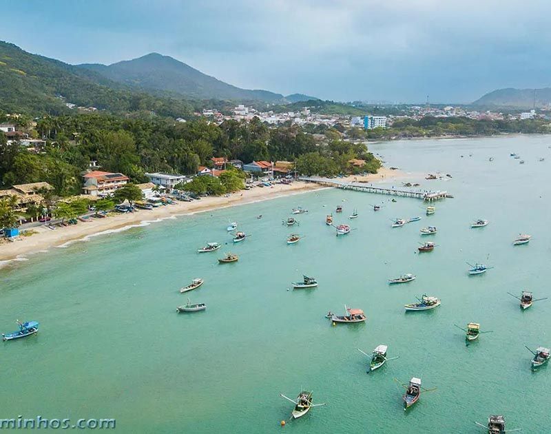 Vista aérea de uma baía tranquila de águas turquesa, pontilhada por inúmeros barcos de pesca pequenos, perto de uma praia de areia e uma encosta verdejante.