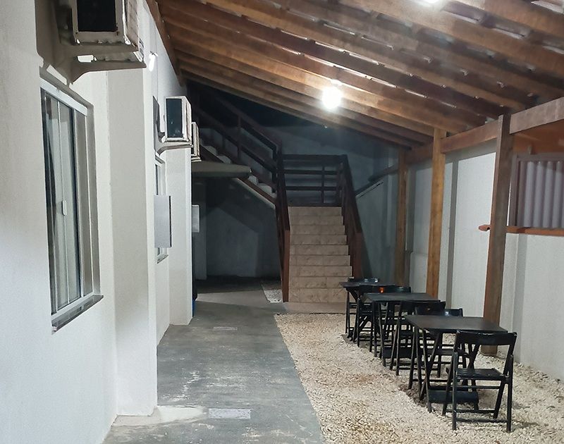 Uma área de refeições ao ar livre com mesas e cadeiras pretas, dispostas sobre cascalho sob um pátio com teto de madeira próximo a uma escada.