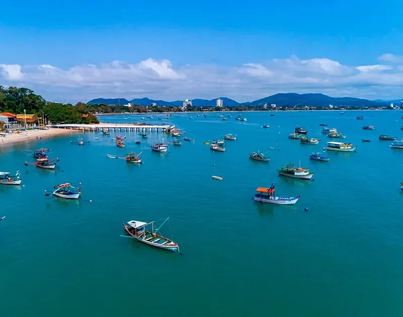 Uma vista panorâmica de inúmeros barcos pequenos ancorados em uma baía tranquila de águas turquesa, sob um céu azul límpido, com montanhas ao longe.