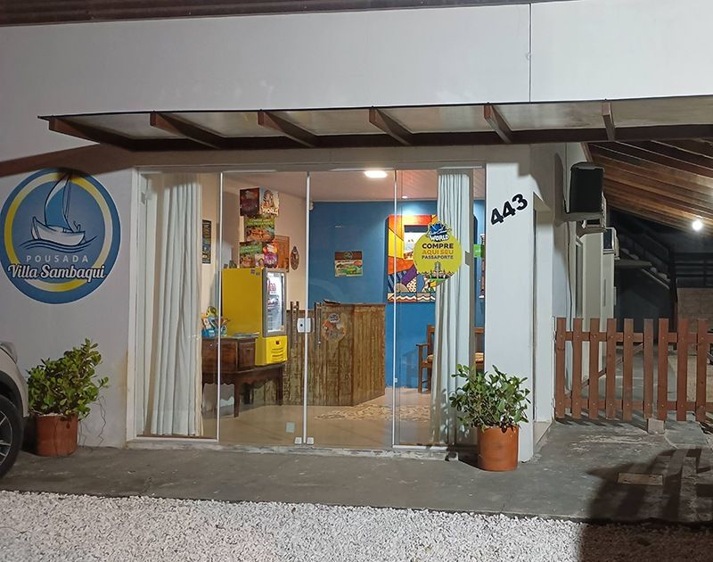 Entrada da Pousada Ilha Sununga à noite, com fachada de vidro, geladeira amarela e vasos de plantas.